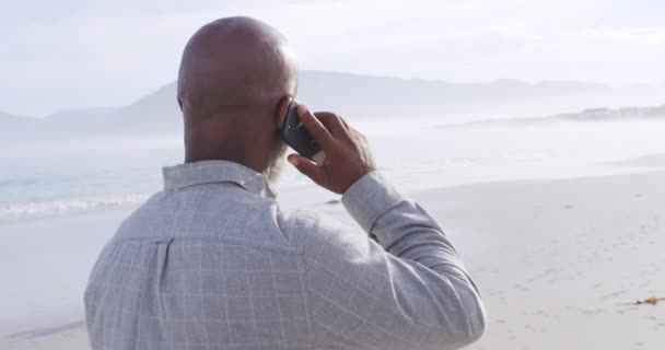成熟した混合レースの男が自分の携帯電話で話し 海のビーチを歩いているの背面ビュークローズアップ — ストック動画
