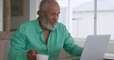 Kahve içen ve evde bir dizüstü bilgisayar kullanarak olgun bir karışık ırk adam yakın ön görünüm