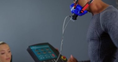 Genç bir Kafkas kadın kişisel eğitmen sonuçları kontrol ederken bir koşu bandı üzerinde çalışan ve eğitim sırasında bir metabolik gaz analizörü kullanarak genç bir karışık ırk adam Yan görünümü