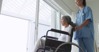 Genç bir karışık ırk kadın hemşire yan görünümü pencereden dışarı bakmak için tekerlekli sandalyede olgun bir karışık ırk kadın iterek