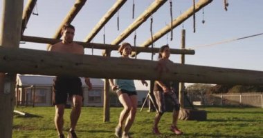 İki beyaz kadın ve beyaz bir genç adamın acemi birliği eğitimi sırasında açık hava spor salonunda bir engelin üzerinden atlarken yan görünüm.