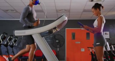 Genç bir karma ırk kadın kişisel eğitmen sonuçları kontrol ederken bir koşu bandı üzerinde çalışan ve eğitim sırasında bir metabolik gaz analizörü kullanarak genç bir karışık ırk adam Yan görünümü