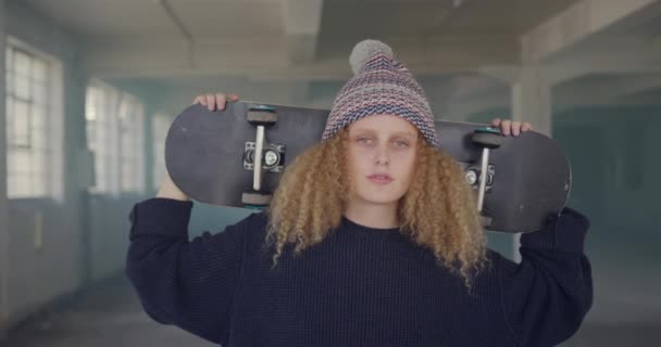 空の倉庫で腰の若い白人女性の正面図 彼の肩にスケートボードを運ぶ — ストック動画
