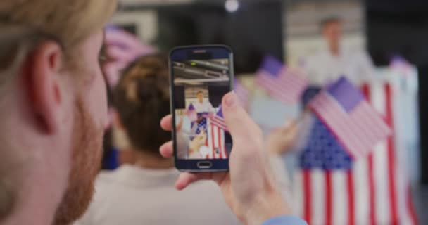 一个拿着智能手机的白人男子站在讲台上 在一个政治集会上挂着美国国旗 观众们挥舞着国旗 — 图库视频影像