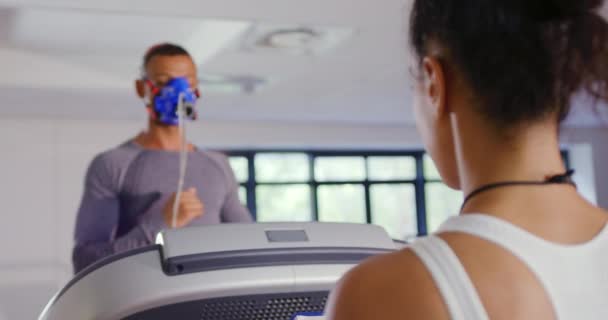 一个年轻的混合种族男子在跑步机上跑步 并在训练期间使用代谢气体分析仪与年轻的混合种族女性私人教练在前景的前视图 — 图库视频影像