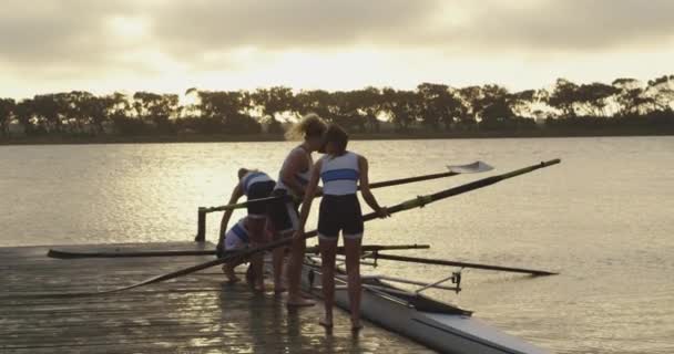 オールを準備する桟橋に立つ4人の若い成人白人女性の漕ぎ手のチームの側面図と川での訓練のためのレースシェル — ストック動画