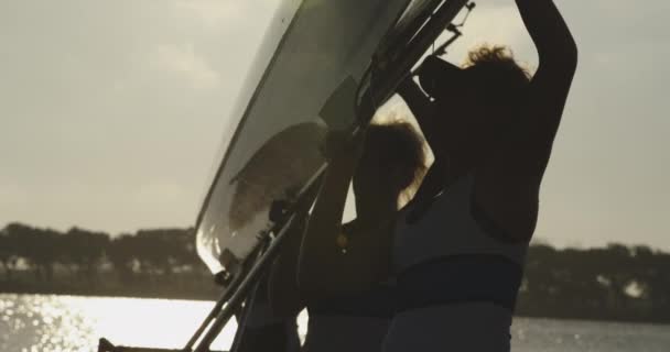 一支由四名剪影的年轻成年白种女性赛艇运动员组成的队伍 站在河边的码头上 举起赛车壳 — 图库视频影像
