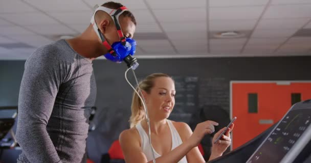 若い白人女性パーソナルトレーナーがタブレットで結果をチェックしている間 トレッドミルで走り トレーニング中に代謝ガス分析器を使用する若い混合レースの男性の側面図 — ストック動画