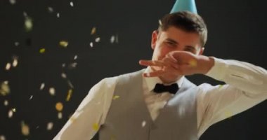 Bir papyon, yelek ve kağıt parti şapkası giyen mutlu bir genç Kafkas adam ön görünümü yakın, bir partide düşen konfeti ve balonlar altında dans, karanlık bir arka plan üzerinde