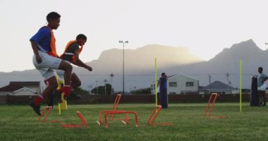 İki yetişkin melez kadın rugby oyuncusu antrenman sırasında önlük giyip engellerden atlarken, arka planda takım arkadaşları vardı.
