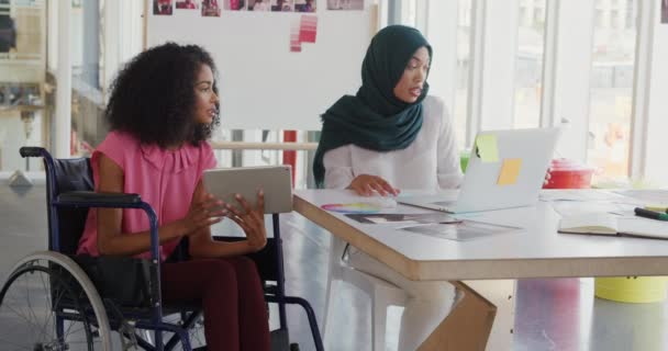 两个年轻的男女混血女企业家一起工作 在一个现代化的办公室里交谈 一个坐在轮椅上 拿着平板电脑 另一个戴头巾的女人则坐在办公桌前 用笔记本电脑 — 图库视频影像
