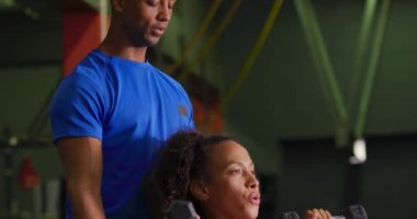 Yan görünüm yakın genç bir karışık ırk erkek kişisel antrenör ile bir spor salonunda egzersiz genç bir karışık Yarış kadın, dumbbells kullanarak
