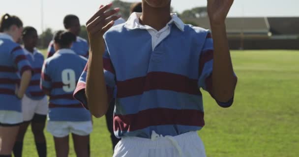一个年轻的男女混合种族橄榄球选手站在橄榄球球场上拿着橄榄球球的画像 把它抛向空中 捕捉它 看着镜头 微笑着 队友们在后台交谈 — 图库视频影像
