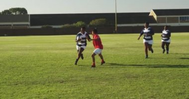 Rugby maçında sahada koşan bir grup yetişkin, etnik çeşitlilikte kadın rugby oyuncusunun ön görüntüsü ve bir oyuncu diğer oyuncuyu bir oyuncuyla yere seriyor.