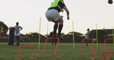 Arka planda takım arkadaşlarıyla antrenman yaparken başlık takan beyaz bir kadın rugby oyuncusunun çeviklik engellerinden atlaması.