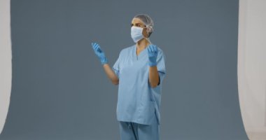 Gri bir arka plan üzerinde önlük, yüz maskesi ve cerrahi eldiven giyen, etrafa bakan genç bir Kafkas kadın cerrah Ön görünümü