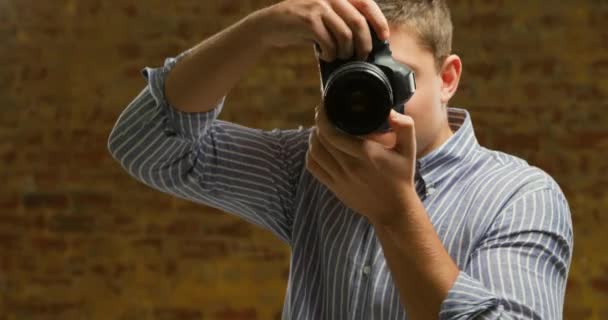 Vorderseite Nahaufnahme eines jungen kaukasischen männlichen Fotografen, der mit einer digitalen Slr-Kamera fotografiert, die Bilder auf der Rückseite der Kamera überprüft und lächelnd vor einer Backsteinmauer steht