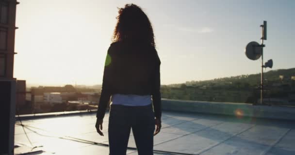 背景に建物を持つ都市の屋上に立つヒップ若い混合人種女性のバックビュー 腕を伸ばし 太陽光でバックライト — ストック動画
