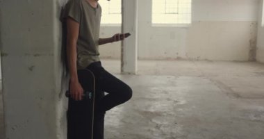 Boş bir depoda bir kalça genç karışık ırk adam Yan görünümü, bir kaykay tutan bir duvara yaslanmış ve bir akıllı telefon kullanarak