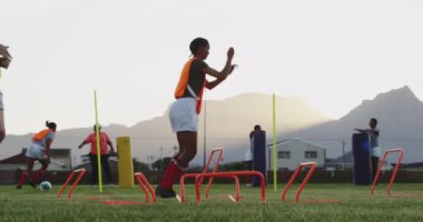 Yetişkin, melez bir kadın rugby oyuncusu ve genç, beyaz bir kadın rugby oyuncusu antrenman sırasında arka planda takım arkadaşlarıyla atlama engelleri takıyor.
