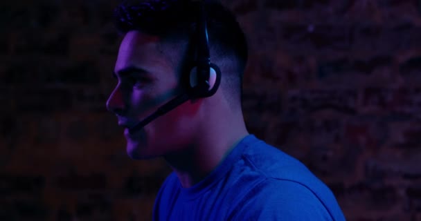 一个年轻的白种人男子在光线昏暗的工作区中用耳机在电脑前工作的侧视图特写 — 图库视频影像