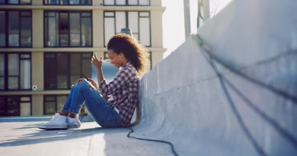 ヒップ若い混合レースの女性のサイドビュークローズアップは 太陽の光でバックライト付き 背景に建物と都市の屋上に座って 彼女のスマートフォンを使用して — ストック動画