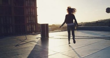 Arka planda binalar ile kentsel bir çatı üzerinde çalışan bir kalça genç karışık ırk kadın Geri görünümü, kollar uzanmış, güneş ışığı ile arkadan aydınlatmalı