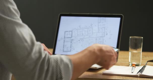 一个年轻的白种人男子坐在办公桌前 使用笔记本电脑拿起智能手机 并使用它 在电脑屏幕上可以看到建筑平面图的侧视图中段 — 图库视频影像