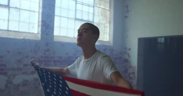 空の倉庫でヒップ若い混合レースの男の正面図 青い煙でアメリカの旗を保持し — ストック動画