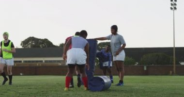 Üç yetişkin, karışık ırk bayan rugby oyuncusunun antrenman sırasında antrenman çantalarıyla antrenman yapmalarına, takım arkadaşlarının ise arka planda antrenman yapmasına dikkat edin.