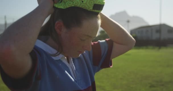 サイドビューアップ若い大人の白人女性ラグビー選手の近くにラグビーピッチに立って 彼女のヘッドガードを固定します — ストック動画