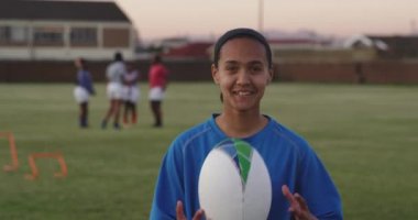 Genç yetişkin bir rugby oyuncusunun portresi. Bir spor sahasında durmuş rugby topunu tutuyor, fırlatıyor, yakalıyor ve kameraya bakıp gülümsüyor. Takım arkadaşları arka planda antrenman yapıyor.