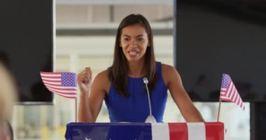 Siyasi bir mitingde seyirciye hitap eden ve yumruğunu kaldıran bizlerin bayraklarıyla süslenmiş bir kürsüde duran gülümseyen genç bir melez kadının ön görüntüsü.