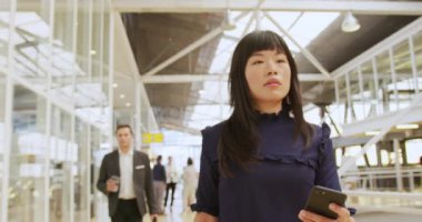 Genç Asyalı bir iş kadınının elinde akıllı bir telefonla modern bir fuayeden iş konferansına yürüdüğü ön manzara. Diğer iş delegeleri arka planda yürürken görülebilir