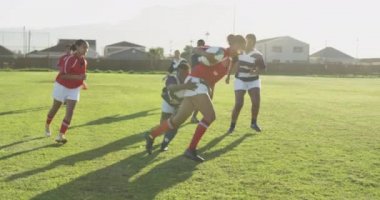Bir ragbi maçında sahada koşan bir grup yetişkin, çok ırklı bayan rugby oyuncusunun yan görüntüsü ve bir oyuncu bir oyuncuyla bir diğerini yere seriyor.