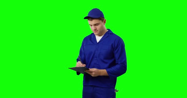 青いオーバーオールとキャップを身に着けている若い白人のハンディマンの正面図クリップボードを保持し 書き込みとカメラ グリーンスクリーンを見て — ストック動画