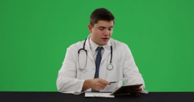 Yeşil bir arka plan üzerinde konuşan ve yazan, tablet kullanan genç beyaz bir erkek doktorun ön görünümü