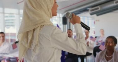 Yan görüntüde elinde mikrofonla ayakta duran genç bir Asyalı kadın var. Siyasi bir kongrede seyirciye sesleniyor. Arka planda izleyiciler bizi dinliyor ve bayrakları tutuyor.