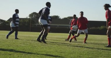Bir ragbi maçı sırasında sahada koşan bir grup yetişkin, çok ırklı kadın rugby oyuncusunun ön görüntüsü.