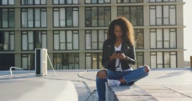 Ön görünüm yakın bir kalça genç karışık ırk kadın oturan ve arka planda bir bina ile kentsel bir çatı üzerinde akıllı telefon kullanarak, güneş ışığı ile arkadan aydınlatmalı