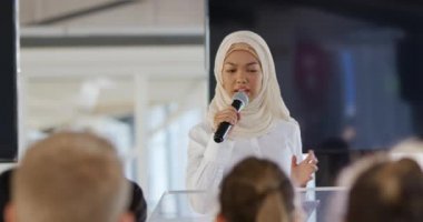Ön planda, kürsüde tesettür takmış genç bir Asyalı iş kadınının bir iş konferansında seyirciye hitap etmek için el kol hareketi yapması ve mikrofon kullanması görülüyor. Seyircilerin kafalarının arkası ön planda görünüyor.