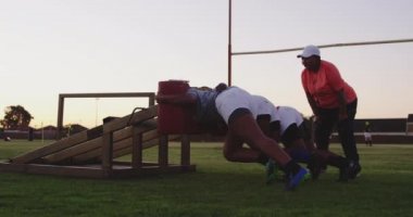 Orta yaşlı melez bir kadın rugby koçunun yan görüntüsü. Bir spor sahasındaki takım antrenmanı sırasında üç yetişkin Afrika kökenli Amerikalı kadın rugby oyuncusuna, hücum makinesi kullanma talimatı veriyor.