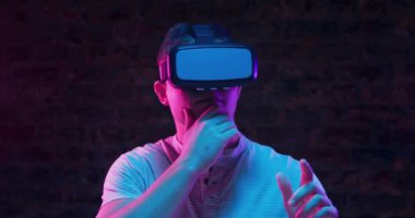 Vr kulaklık takan ve etrafa bakan genç bir Kafkas adamın ön görünümü, bir eliyle çenesine, diğer eliyle sivri parmakla önünde, siyah bir arka plan üzerinde pembe ve mavi ışıkla aydınlatılmış,