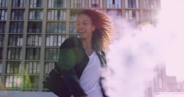 Bir kalça genç karışık ırk kadın gülümseyen ve arka planda bir bina ile kentsel bir çatı üzerinde sis bombası kullanarak, güneş ışığı ile arkadan aydınlatmalı ön görünümü