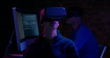 Vr kulaklık takan genç bir karma yarış kadını yakın, etrafa bakıyor ve karanlık aydınlatılmış bir çalışma alanında ellerini hareket ettiriyor, arka planda beyaz bir adam da vr kulaklık takan bir bilgisayarda çalışıyor