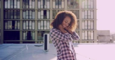 Arka planda binaları ile kentsel bir çatı üzerinde duran bir kalça genç karışık ırk kadın Ön görünümü, kamera bakarak, güneş ışığı ile arkadan aydınlatmalı