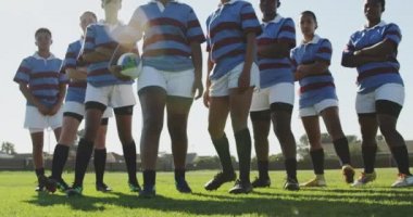 Rugby sahasında dizilmiş, kolları çaprazlanmış, ragbi sahasında kameraya bakan genç, çok ırklı bayan rugby oyuncularından oluşan bir takımın portresi. Öndeki kadın bir ragbi topu tutuyor.