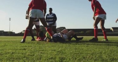 Rugby maçında sahada koşan bir grup yetişkin, etnik çeşitlilikte kadın rugby oyuncusunun ön görüntüsü ve bir oyuncu diğer oyuncuyu bir oyuncuyla yere seriyor.