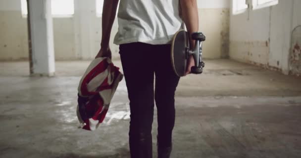 空の倉庫でヒップ若い混合レースの男の背面図 歩くと歩いて アメリカの旗とスケートボードを運ぶ — ストック動画