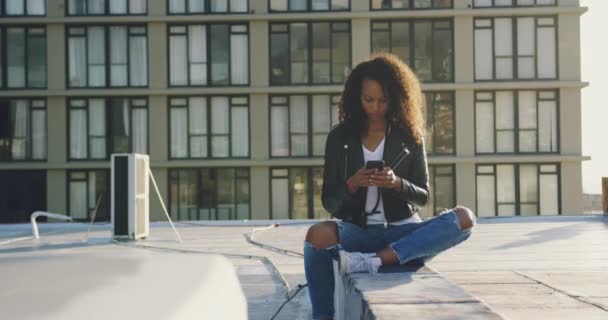前视图特写一个时髦的年轻混血女人坐在城市屋顶上 用她的智能手机 背景是一座建筑物 被阳光背光 — 图库视频影像